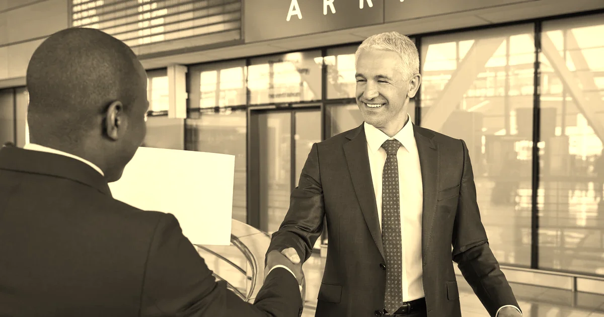 Chauffeur en costume sombre accueillant un client d’affaires au panneau nominatif dans une zone arrivées d’aéroport, traitement sépia presse B2B