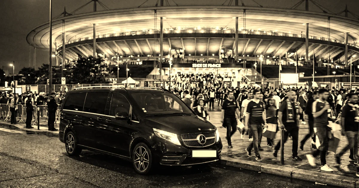 Van de grande remise à l’arrêt le long du trottoir devant le Stade de France illuminé, foule de spectateurs quittant l’enceinte à la nuit tombée, traitement sépia presse B2B